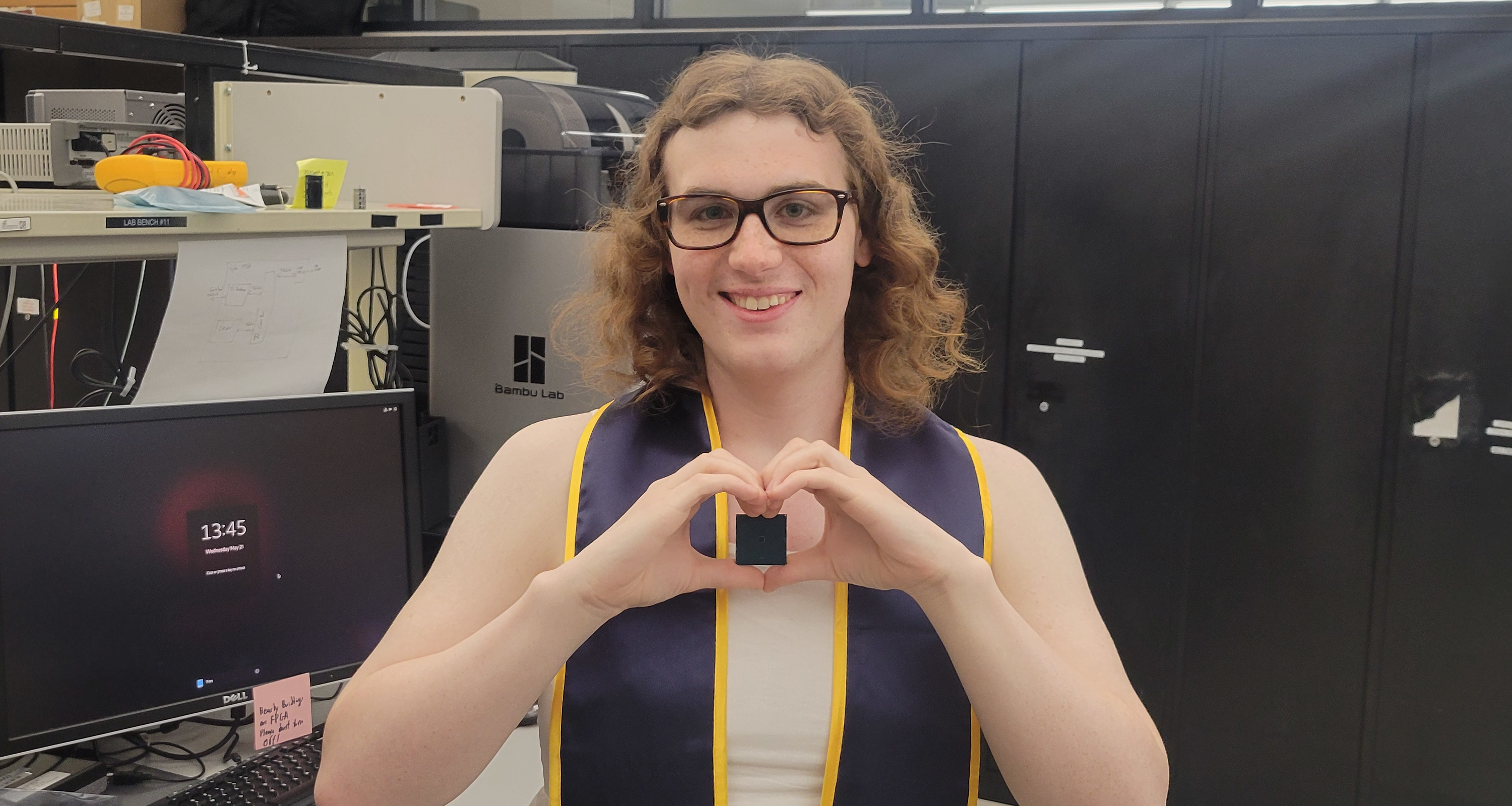 Picture of Jasmine in graduation attire inside the BWRC lab holding the Spring 2024 class chip with hands cupped in a heart shape